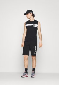 Svart ärmlös skjorta med vit horisontell rand och "REPLAY"-branding, matchad med svarta shorts med "ALPHA"-text. Grå sneakers.