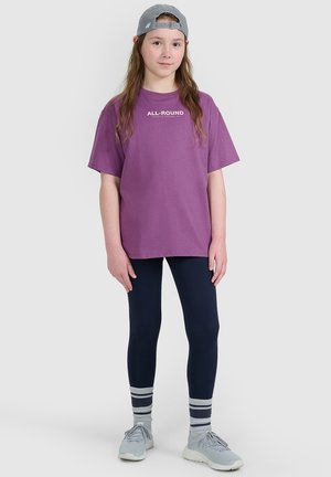 Młoda osoba ubrana w szarą czapkę, fioletowy t-shirt z napisem „ALL-ROUND”, granatowe legginsy, paskowane skarpetki i szare trampki stoi na białym tle.