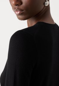 Gros plan d'une personne portant un pull noir et de grandes boucles d'oreilles rondes argentées réfléchissantes, montrant le bas du visage, le cou et l'épaule sur un fond uni.