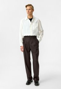 Witte lange mouwen shirt met zak, gecombineerd met high-waisted donkerbruine broeken en zwarte schoenen. Soepele textuur en relaxte pasvorm.