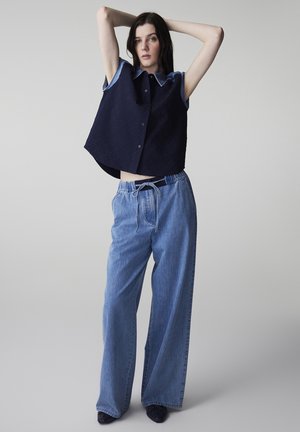 Woman standing with arms raised wearing a sleeveless navy buttoned top and wide-leg light blue denim pants with drawstring waist.