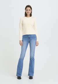 Cardigan crème à manches longues avec un col rond, fermeture à boutons à l'avant, assorti à un jean évasé bleu clair et des bottines noires.