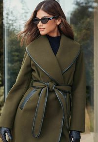 Alyvuogių spalvos paltas su apvyniojimo dizainu, su juodais odiniais akcentais ir dirželiu ties liemeniu. Textūruotas audinys ir platūs apykaklės išbaigia įvaizdį.