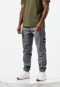 Grå cargo bukser med sidelommer, laget av et glatt stoff, kombinert med en grønn t-skjorte og hvite sneakers på en marmoroverflate.