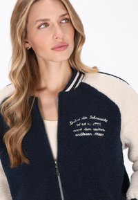 Marineblå og cremefarvet farveblokeret bomberjakke lavet af blødt, struktureret stof; har broderet tekst og lynlås foran med stribede kraveaccenter.