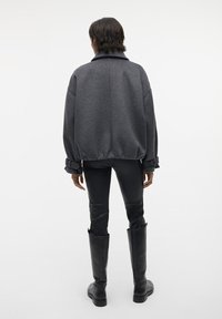 Giacca in pile grigia oversize con colletto, orlo e polsini elastici, abbinata a pantaloni aderenti neri lucidi e stivali neri al ginocchio.