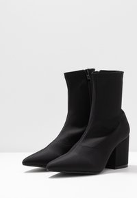 Bottines noires en tissu lisse et extensible, avec un bout pointu et un talon bloc. Dotées d'une fermeture éclair sur le côté pour un enfilage facile.