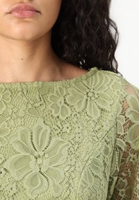 Haut en dentelle dans un vert atténué, avec des motifs floraux, un large décolleté et des manches transparentes. Le matériau a une finition délicate et texturée.