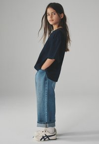 Ragazza giovane con lunghi capelli castani, indossa una maglietta nera larga, jeans blu risvoltati, calze bianche e sneaker bianche con strisce nere, in piedi di profilo.