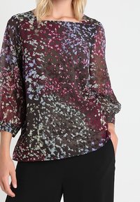 Blouse transparente avec un col rond plongeant, arborant un imprimé floral foncé aux nuances de violet, bleu et vert. Manches longues bouffantes en tissu texturé.