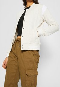Jasnoszara bomberka na zamek z zatrzaskami, ribbowanymi mankietami i białymi akcentami na ramionach, w połączeniu z brązowymi spodniami cargo.