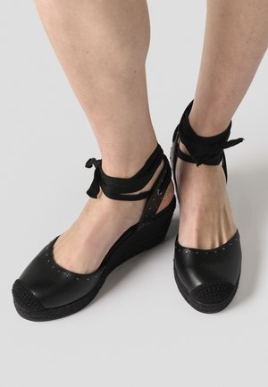 Pėdos juodais pusiau platforminiais espadriliais su vinutėmis įsegtais pirštų gaubtais ir juodomis čiurnos raištelėmis ant šviesiai pilko fono.