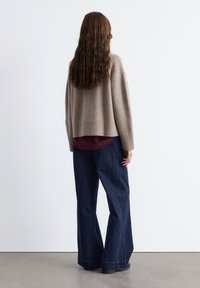 Beige gestrickter Pullover mit lockerer Passform und geripptem Saum, über einem burgunderfarbenen Oberteil getragen. Dunkelblaue weite Jeans und schwarze Schuhe.