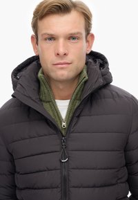 Sort pufferjakke med hætte, med en lynlåslukning foran og quiltet tekstur. Laget over en grøn ribbet sweater.