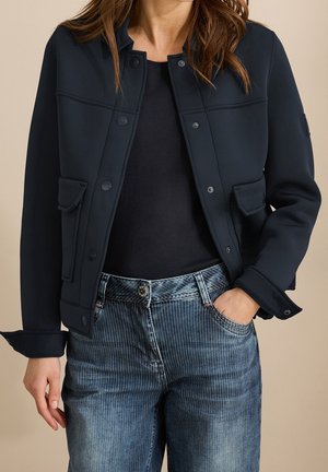 Femme portant une veste noire boutonnée sur un top noir, avec une main dans la poche d'un jean bleu à fines rayures, sur un fond neutre.