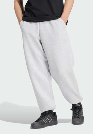 Osoba nosząca jasnoszare luźne spodnie dresowe i czarne buty sportowe Adidas stoi ze złożonymi rękami w kieszeniach.