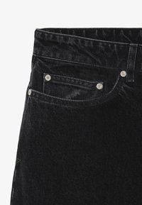 Jean de denim negro con una cintura alta, diseño de cinco bolsillos, textura sutil y acentos de herrajes metálicos. Incluye detalles de costura distintivos.
