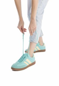 Zapatillas deportivas de color azul claro con rayas beige, parte superior de textil y suela de goma, atándose mientras usa unos jeans de mezclilla claros.