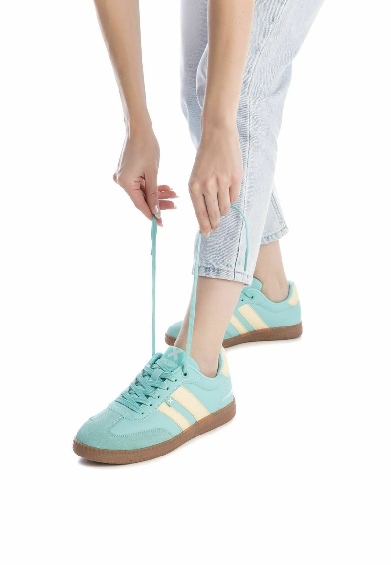 Zapatillas deportivas de color azul claro con rayas beige, parte superior de textil y suela de goma, atándose mientras usa unos jeans de mezclilla claros.