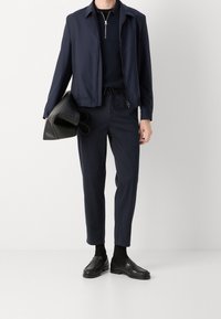 Mörkblå jacka med ställkrage och dragkedja, tillsammans med mörkblå byxor och svarta loafers. Personen håller en svart läderväska.