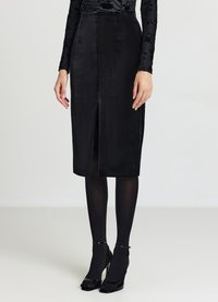 Schwarzer Bleistiftrock aus Samt mit Frontschlitz, hoher Taille und strukturierter Form. Kombiniert mit transparenten schwarzen Strumpfhosen und spitz zulaufenden Schuhen.