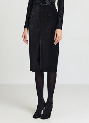 Gonna a matita in velluto nero con spacco frontale, vita alta e design strutturato. Abbinata a collant neri trasparenti e décolleté a punta.