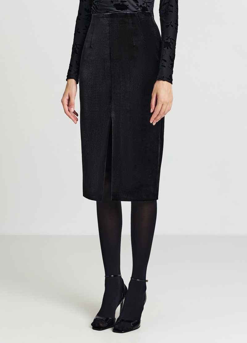 Schwarzer Bleistiftrock aus Samt mit Frontschlitz, hoher Taille und strukturierter Form. Kombiniert mit transparenten schwarzen Strumpfhosen und spitz zulaufenden Schuhen.