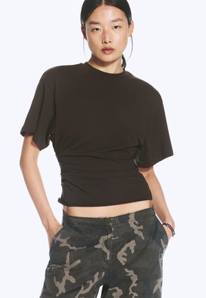 Jeune femme aux cheveux noirs attachés, portant un haut noir moulant à manches courtes et un pantalon camouflage foncé, les mains dans les poches.