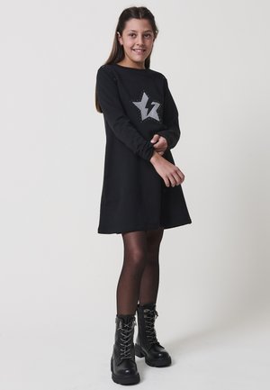 Niña pequeña vestida con un vestido negro con diseño de estrellas y rayos, medias negras y botas negras con cordones, de pie frente a un fondo liso.