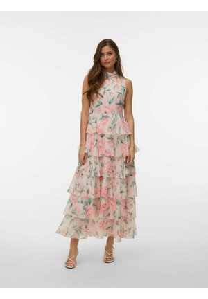 Kobieta o długich włosach, ubrana w bezrękawnikową, warstwową sukienkę z falbankami w różowo-zielony kwiatowy wzór, stoi o neutralnym wyrazie twarzy i białych szpilkach.