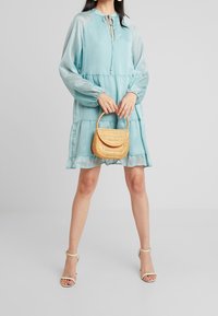 Robe bleu clair à manches longues avec un motif texturé, col à cordon, jupe à volants et un sac à main assorti en tissage. Chaussures à talons avec brides.