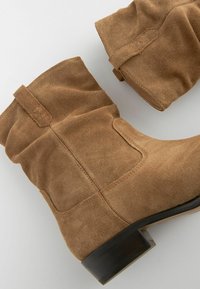 Bottines en suède marron avec une texture douce, un design décontracté et un petit talon empilé. Coutures latérales et un détail de boucle subtil en haut.
