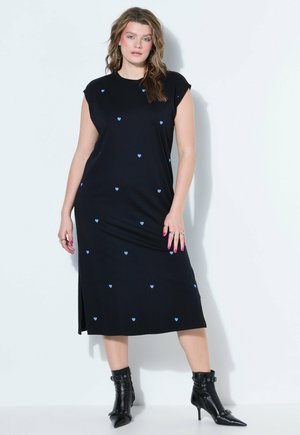 Vrouw staand, gekleed in een zwarte midi-jurk met kleine blauwe hartjes, gecombineerd met zwarte puntige enkellaarzen met gespen.