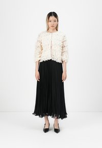 Giacca texturizzata avorio con design a ruches, abbinata a una gonna plissettata nera. Scarpe nere a punta completano l'outfit.