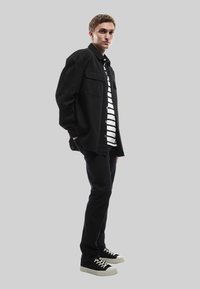 Chaqueta negra de gran tamaño con botones sobre una camisa a rayas, combinada con pantalones negros y zapatillas negras con suelas blancas. Diseño simple y moderno.