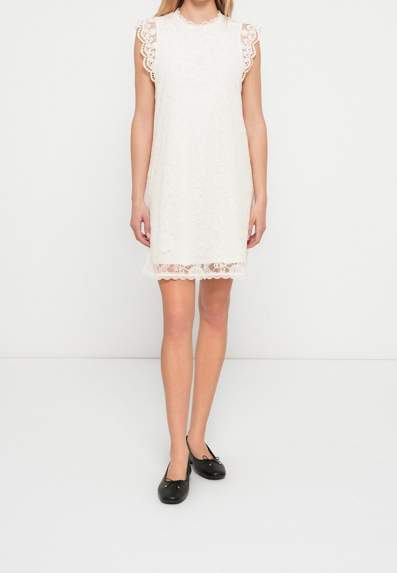 Femme portant une robe en dentelle blanche sans manches avec des bords festonnés et des ballerines noires, debout sur un fond blanc uni.