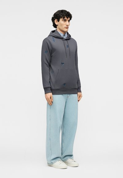 Νεαρός άνδρας με σγουρά σκουρόχρωμα μαλλιά, που φοράει σκούρα γκρι φούτερ με κουκούλα και μπλε κεντημένα σχέδια, ανοιχτό μπλε παντελόνι φαρδιάς γραμμής και λευκά αθλητικά παπούτσια.