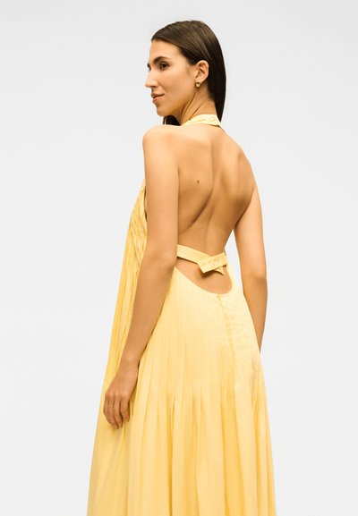 Γυναίκα με μακριά σκούρα μαλλιά που φοράει κίτρινο πλισέ φόρεμα χωρίς πλάτη με λουράκι στο λαιμό και λεπτομέρεια φιόγκου, στραμμένη πλευρικά σε λευκό φόντο.