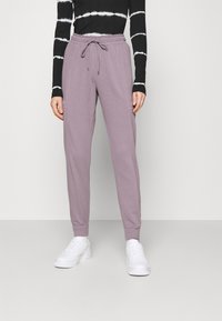 Pantaloni tuta viola chiaro con vita elastica, cordoncino e tasche laterali. Abbinati a sneakers bianche e una maglietta a maniche lunghe a righe nere.