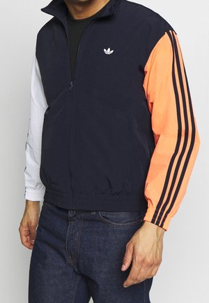 Homme portant une veste de survêtement Adidas zippée avec un corps bleu marine, une manche gauche blanche et une manche droite orange avec des rayures noires.