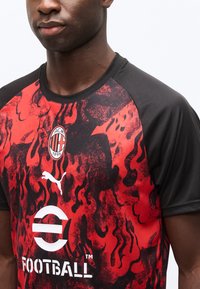 Άνδρας που φοράει κόκκινη και μαύρη φανέλα ποδοσφαίρου της AC Milan με αφηρημένα φλόγινα μοτίβα και λογότυπο Puma στο στήθος.