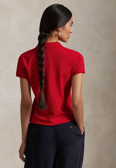 Kvinde med langt fletninghår iført en rød kortærmet polo og navybukser, stående med hænderne i lommerne, vendt sidelæns.