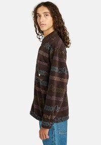 Giovane con lunghi capelli ricci che indossa una camicia di pile a motivi scuri e jeans blu, in piedi di lato contro uno sfondo neutro.