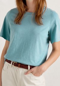 Light blue short-sleeve t-shirt made of soft fabric, paired with a braided brown belt over white pants. Simple and casual design.