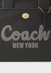 Mustalla tekstuurilla varustettu nahka-tote, jossa on kohokuvioitu tummanharmaa "Coach"-logo ja "New York"-teksti, jota täydentävät kultaiset metalliosat soljessa ja nimilapussa.