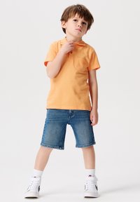 Oranje polo shirt met een borstzak, gecombineerd met denim shorts. Het kind draagt witte hoge sneakers en sokken, staande met de handen op de heupen.
