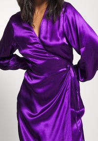 Lila Satinwickelkleid mit langen Ärmeln, V-Ausschnitt und einer Schleife in der Taille. Der Stoff hat eine glatte, glänzende Textur.