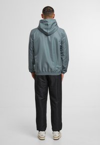 Světle šedá mikina s kapucí a elastickými manžetami, nošená přes černé kalhoty. Vyznačuje se hladkým povrchem a volným střihem.