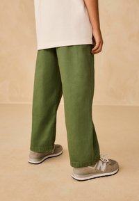 Světle zelené kalhoty širokého střihu s hladkým povrchem, spárované s šedými teniskami New Balance s bílými akcenty a tkaničkami.