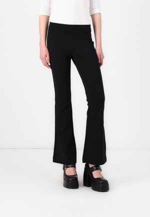 Pantalons évasés noirs en tissu extensible avec une taille haute, assortis à des chaussures plates-formes noires chunky. Texture lisse, design simple.